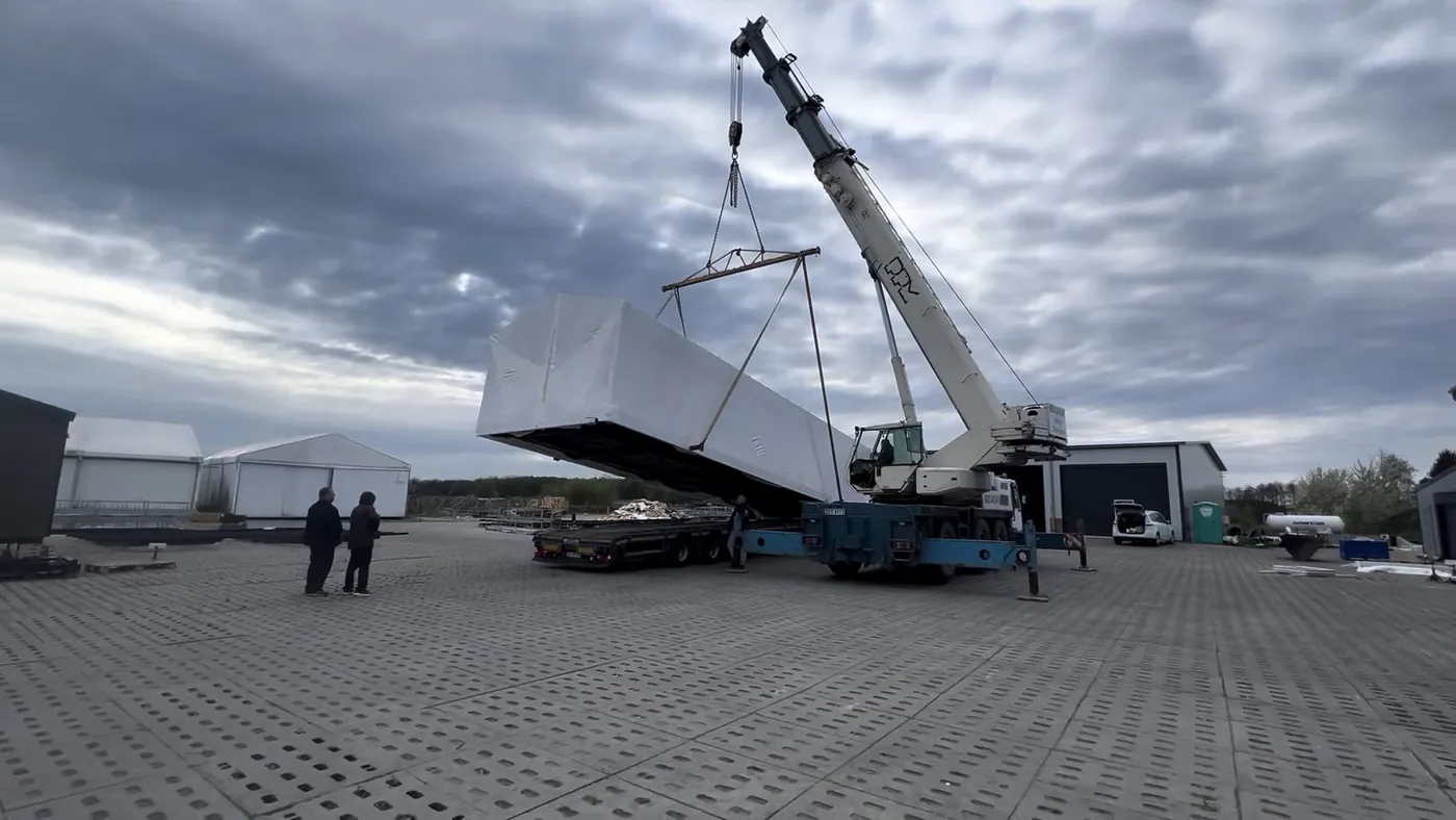 Small house packed in white film, lifted by a mobile crane in a yard, two people standing nearby