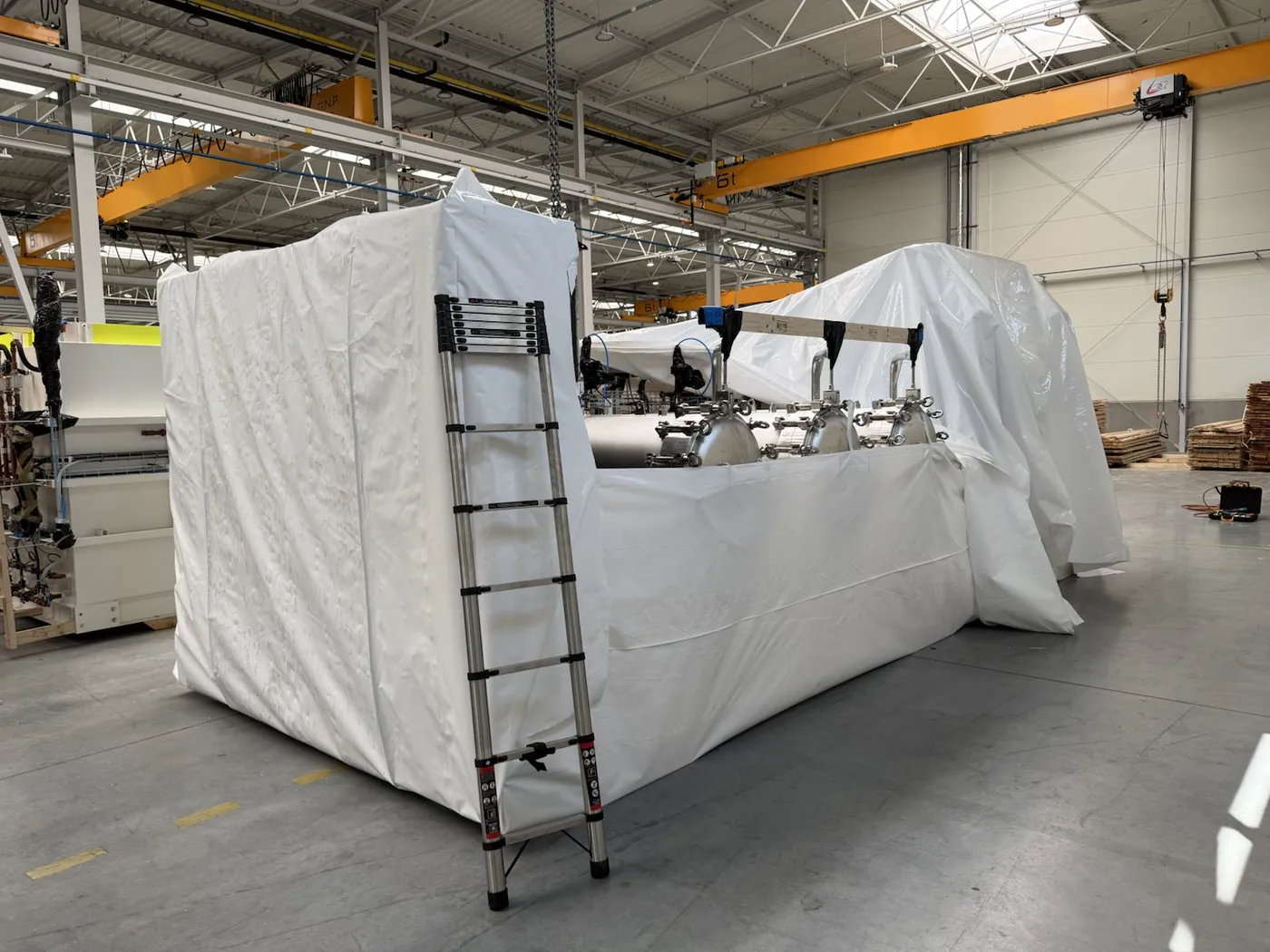 Large machine being wrapped in white shrink wrap film in a production hall, a ladder beside it, an overhead crane in the background