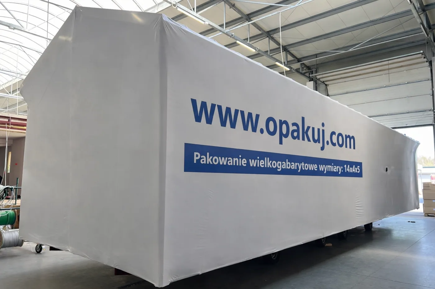 Obiekt o wymiarach 14 na 4 na 5 metrów owinięty białą folią z nadrukiem adresu opakuj.com