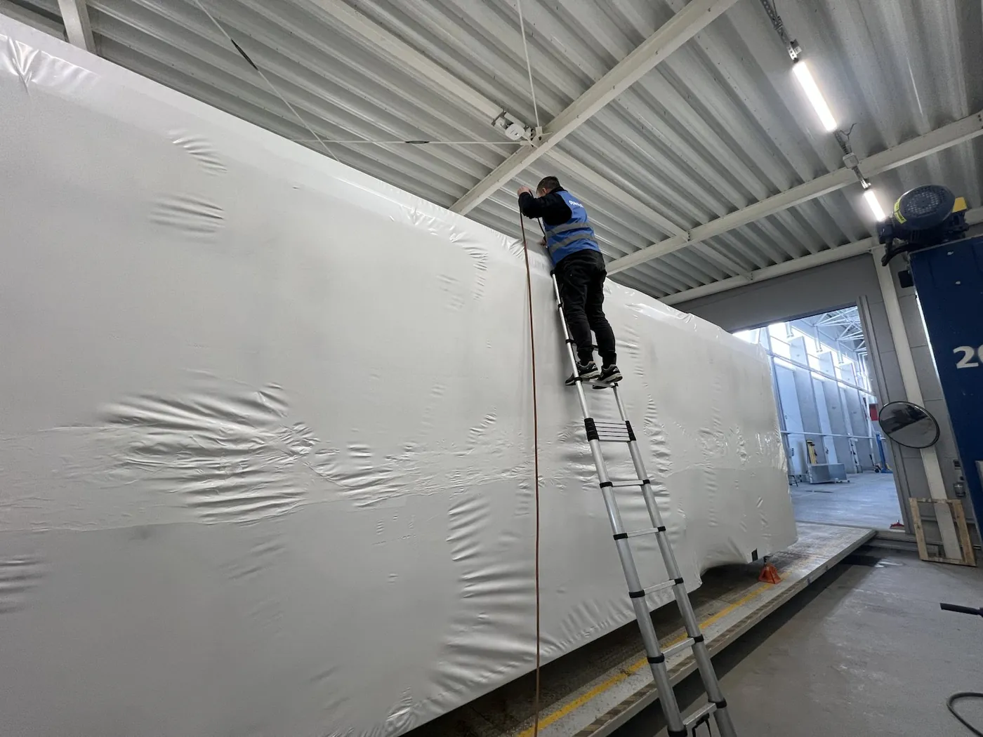 Long frame wrapped in white shrink wrap film in a hall, a worker on a ladder at its top edge