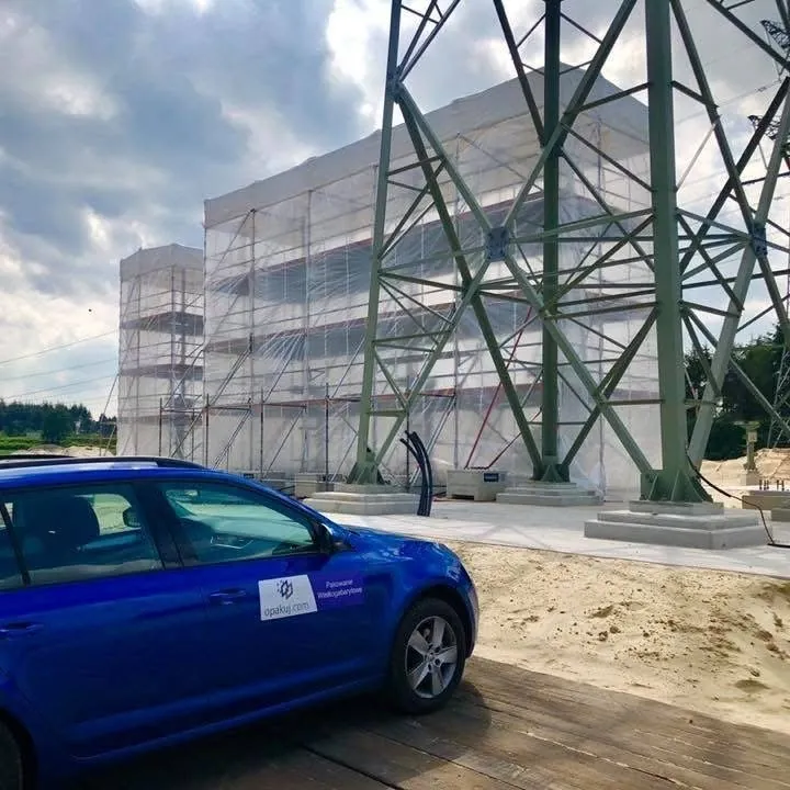 Scaffolding by a building enclosed in white shrink wrap film, a steel power pylon beside it, a car in the foreground