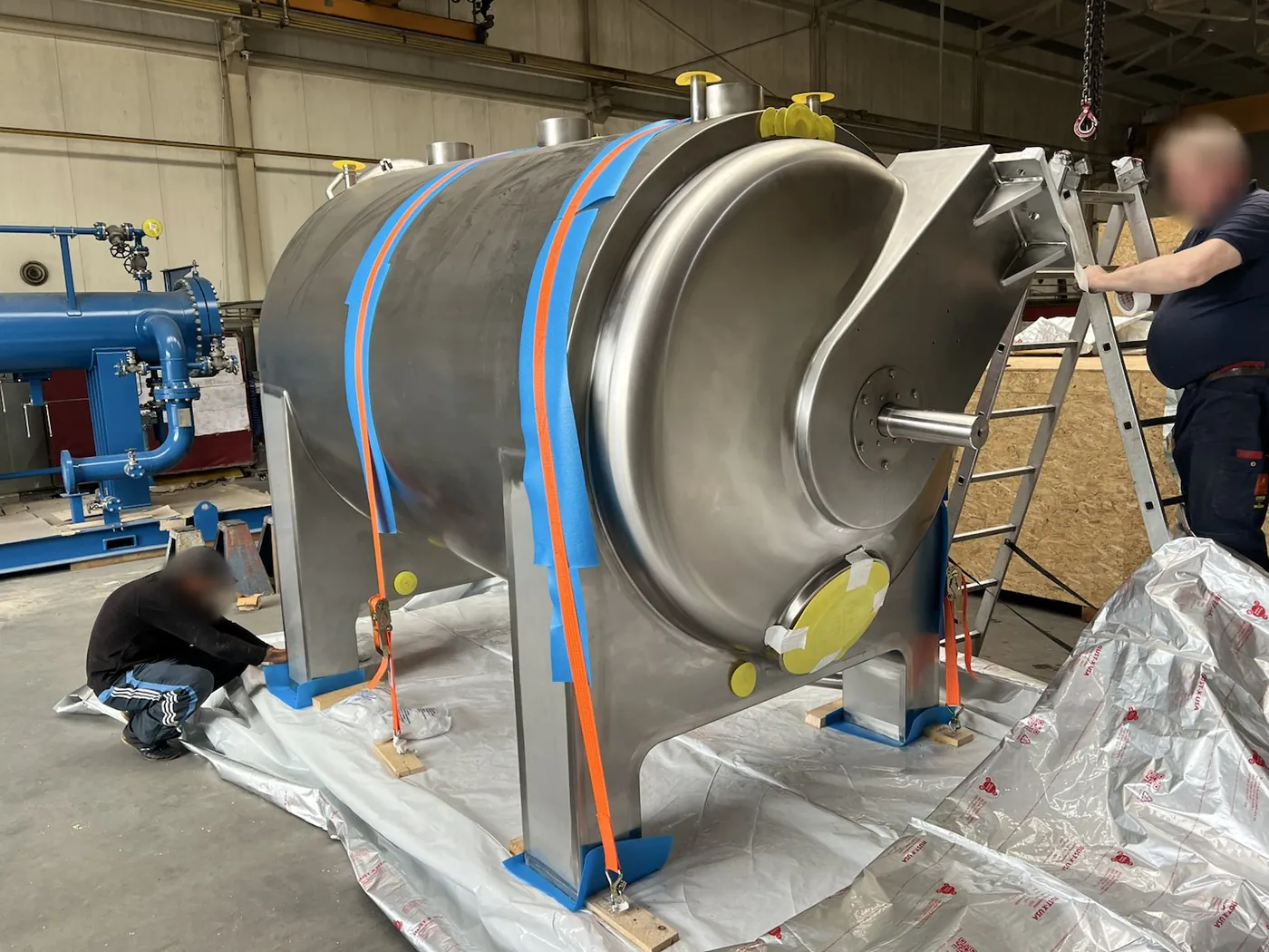 Steel silo on a frame, secured with orange straps, silver barrier foil spread out beneath it, two workers preparing the packing in a hall